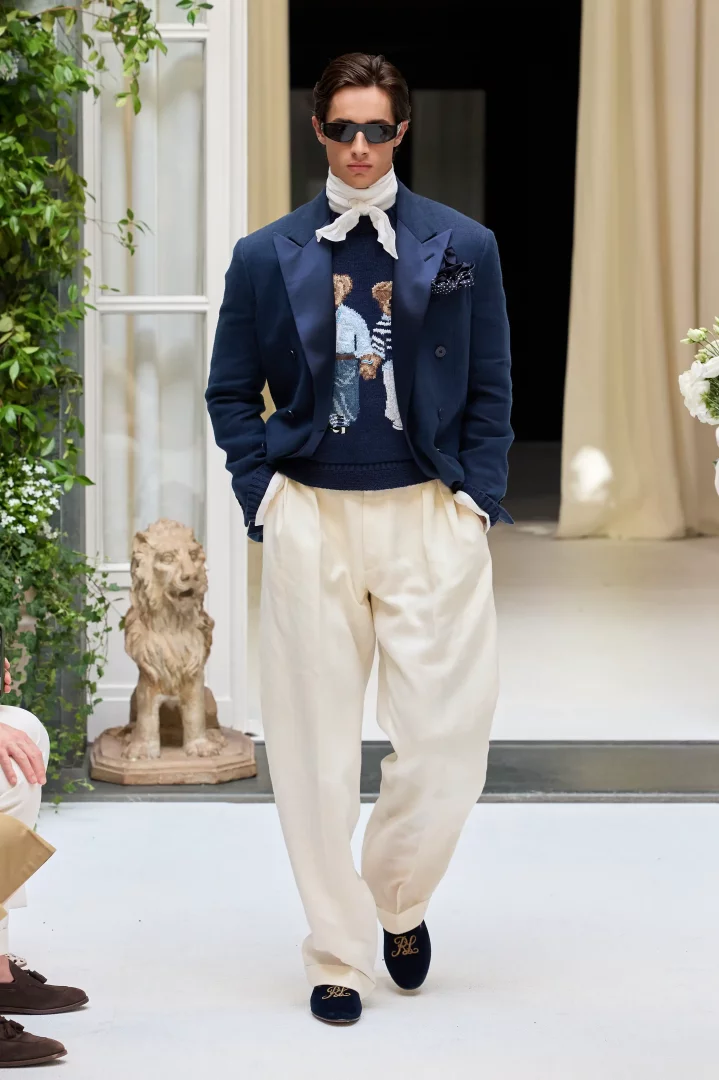 Ralph Lauren'in İlkbahar-Yaz 2027 defilesinden, gözlüklü bir erkek model lacivert kruvaze blazer ceket, Polo Bear desenli lacivert süveter, bol kesim krem pantolon ve logolu lacivert makosenlerle podyumda yürüyerek yeni Amerikan zarafetini yansıtan özgün bir stil sunuyor.