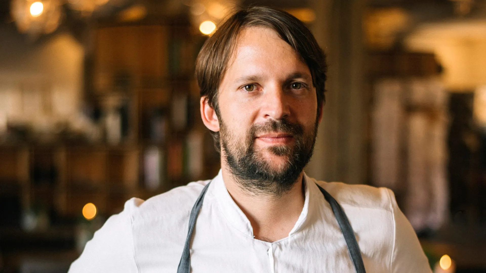 Dünyaca ünlü şef René Redzepi, arkasında hafifçe bulanıklaşmış sıcak ışıklı ve ahşap tonlarında bir restoran ortamında, sade beyaz şef ceketini ve koyu renk önlüğünü giymiş olarak doğrudan kameraya bakıyor, yüzünde sakin bir ifade var.