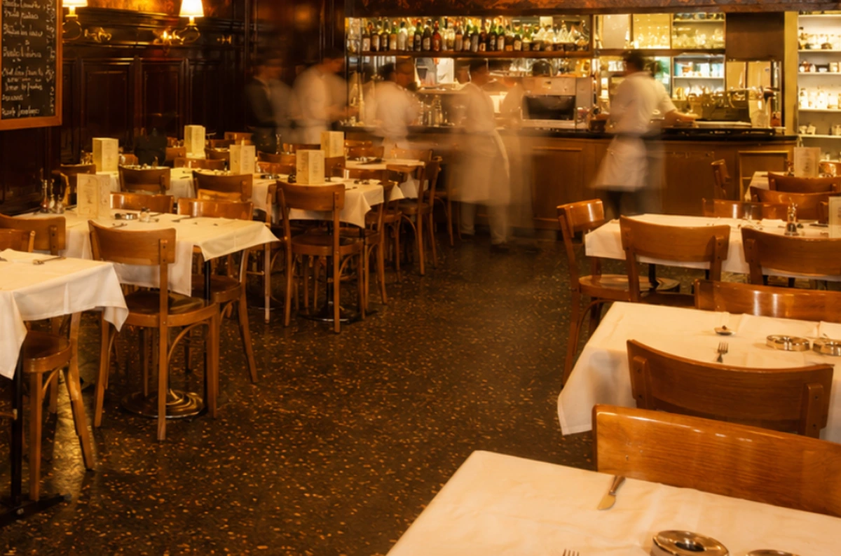 Klasik bir restaurant iç mekanında, beyaz masa örtülü masalar ve ahşap sandalyeler düzenli bir şekilde sıralanmış, arka plandaki bar tezgahında hareketli çalışanlar bulanık bir şekilde görülüyor ve mekanın sıcak, davetkar atmosferini yansıtıyor.