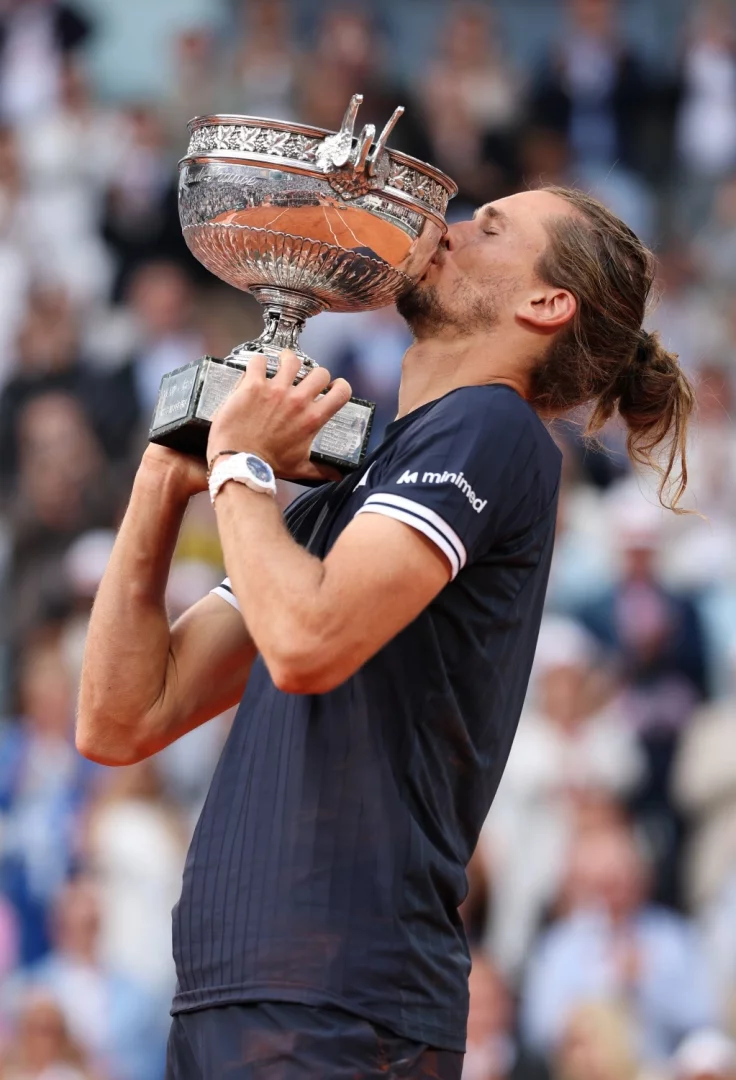 Roland Garros Fransa Açık şampiyonu Alexander Zverev, galibiyetini kutlarken uzun saçları topuz yapılmış, lacivert tişörtü ve kolundaki beyaz Jacob & Co saatiyle gümüş şampiyonluk kupasını coşkuyla öpüyor.