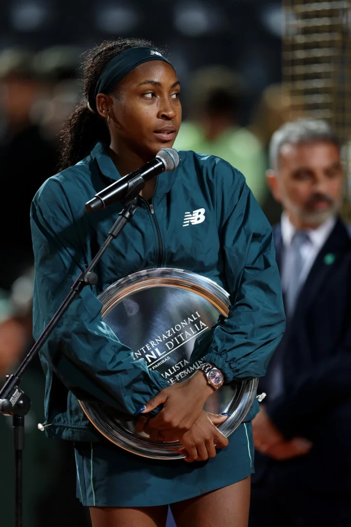 Genç tenisçi Coco Gauff, New Balance eşofmanıyla sahneye çıkmış, Internazionali d'Italia ödülünü iki eliyle kavramış, sol bileğinde mor kadranlı dikkat çekici bir Rolex Oyster Perpetual saat taşıyarak bir mikrofona doğru konuşmak üzere bakıyor.