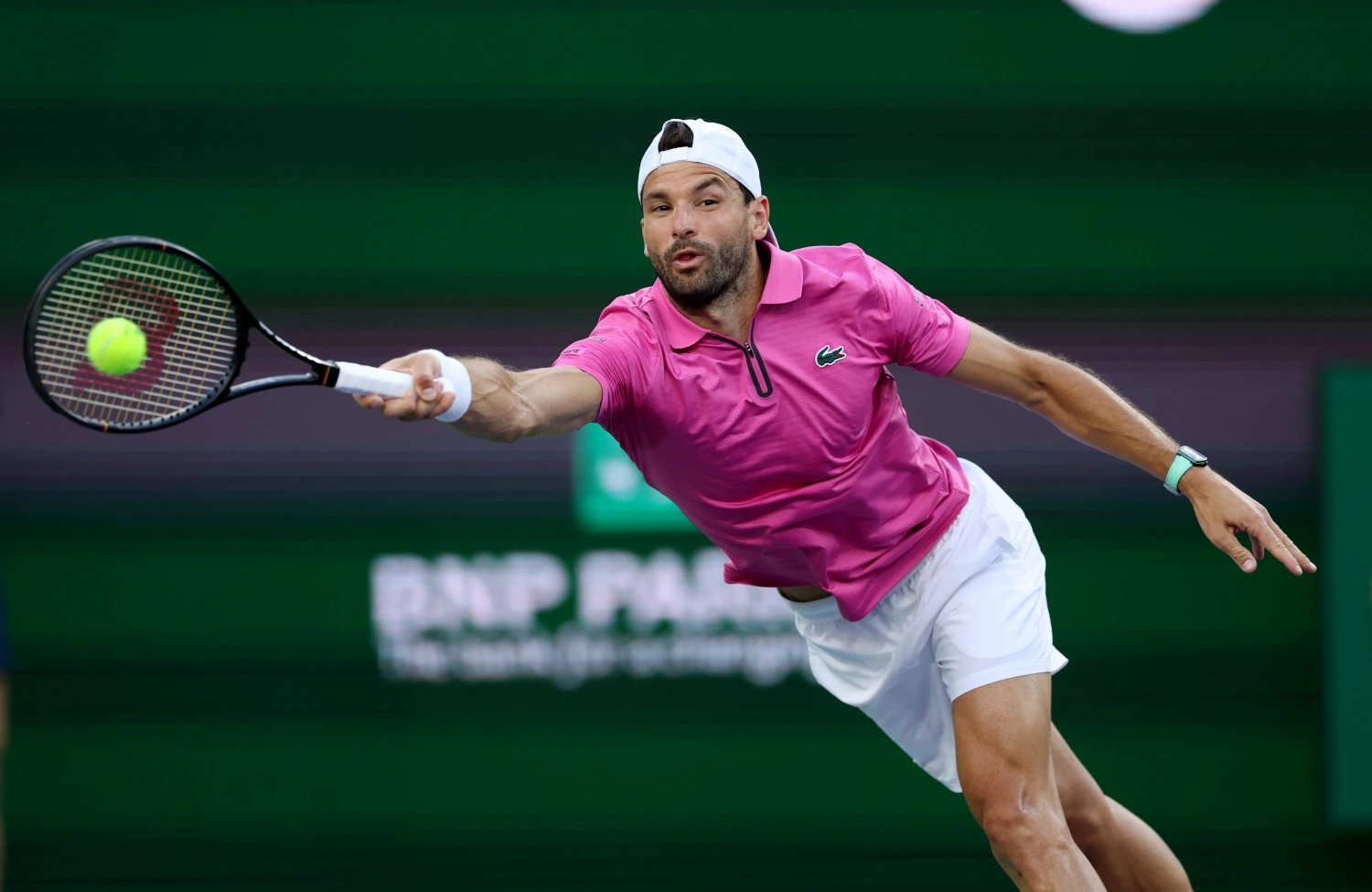 Roland Garros Fransız Açık'ta oynayan Grigor Dimitrov, pembe Lacoste tişörtü, beyaz şortu ve şapkasıyla yeşil kortta tenis topuna uzanırken, bileğindeki modern kol saati net bir şekilde seçiliyor ve arkasındaki bulanık sponsor pankartı maç ortamını tamamlıyor.