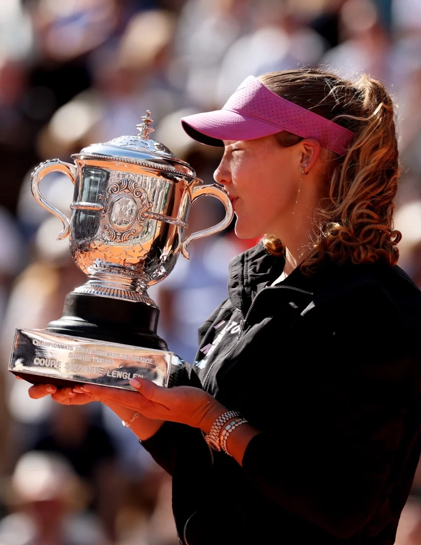 Bir tenisçi, mor siperlikli şapkası ve siyah fermuarlı ceketiyle, dudaklarını öpüyormuşçasına yaklaştırdığı parlak ve gösterişli Fransız Açık Suzanne Lenglen Kupası'nı iki eliyle tutarken görülüyor; bileğindeki taşlarla süslü değerli saat ise belirgin bir şekilde dikkat çekiyor.