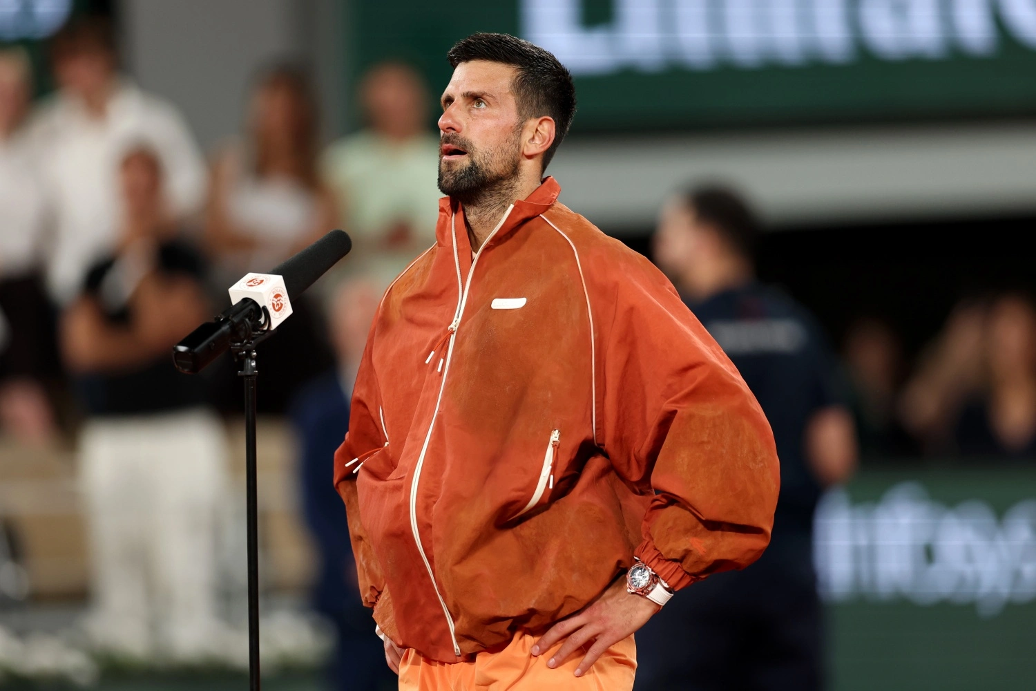 Turuncu renkte sportif bir ceket giyen ve ellerini beline koymuş Novak Djokovic, Roland Garros kortunda podyumda yukarıya doğru dalgınca bakarken sol bileğindeki beyaz kayışlı Hublot Big Bang Unico saatini sergiliyor; yanında mikrofon ve bulanık seyirci kalabalığı görülüyor.