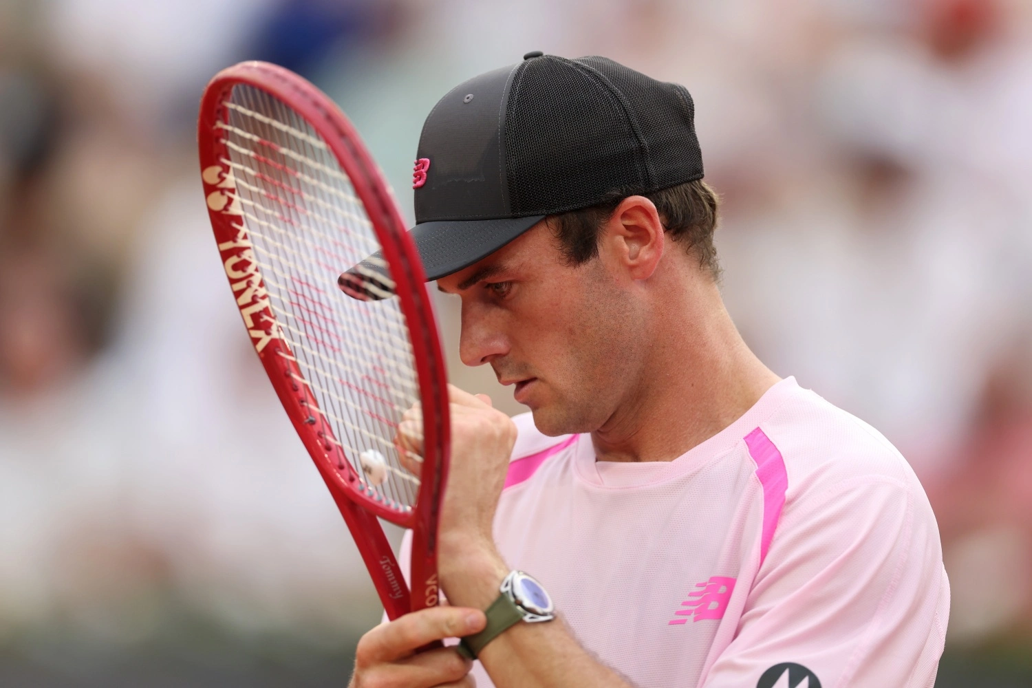 Roland Garros'ta müsabaka anında kırmızı Yonex raketiyle yavaşça ilgilenen, siyah şapkalı ve pembe tişörtlü tenisçi Tommy Paul'un kolunda, göz alıcı kadranı ve zeytin yeşili kayışıyla De Bethune DB28 Steel Wheels lüks kol saati net bir şekilde görülüyor.
