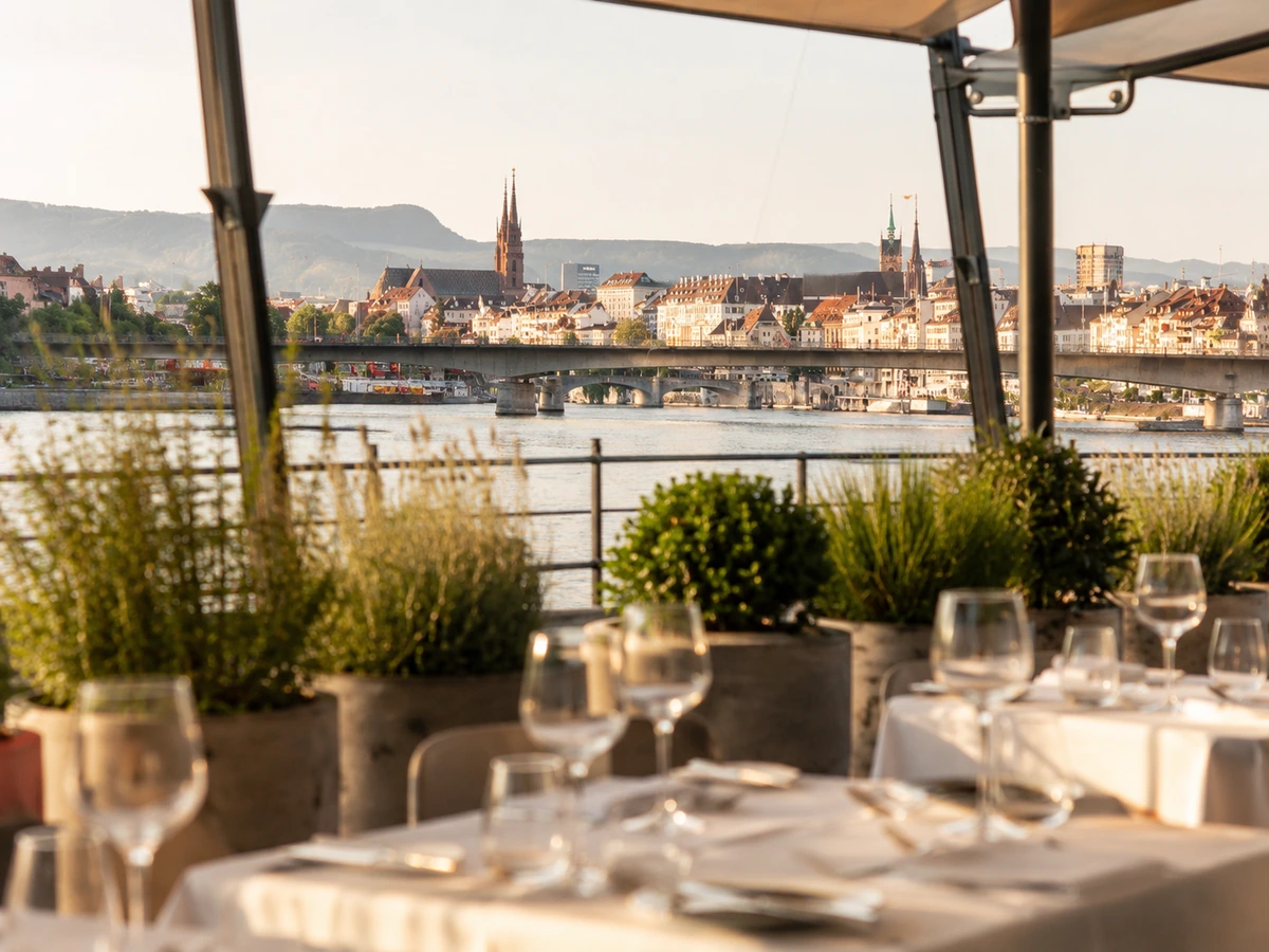 Güneş batarken, Basel şehrinin Ren Nehri üzerindeki köprüler ve tarihi binalarla bezeli panoramik teras manzarasında, şık kadeh ve tabaklarla özenle hazırlanmış masalar bitkilerin arasında konumlanmış.