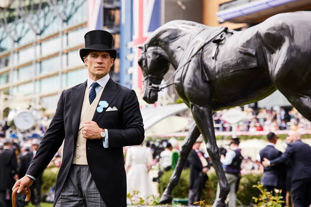 Siyah silindir şapkası, frakı ve çizgili pantolonuyla zarif bir görünüm sergileyen Henry Cavill, Longines sponsorluğundaki Royal Ascot etkinliğinde, kolunda saati ve elinde bastonuyla kameraya bakarken koyu renk bir at heykelinin yanında poz veriyor.