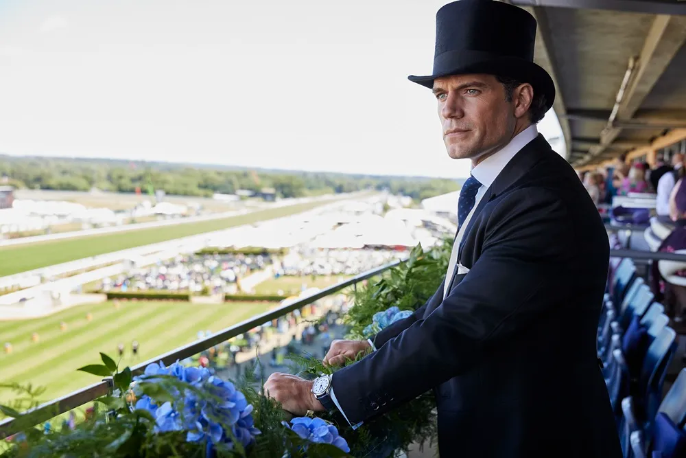 Henry Cavill, Royal Ascot etkinliğinde, mavi çiçekler ve yeşil bitkilerle süslü bir balkonda siyah silindir şapkası ve takım elbisesiyle, sol bileğindeki Longines saatiyle kalabalık at yarışı pistine doğru düşünceli bir şekilde bakarken görülüyor.
