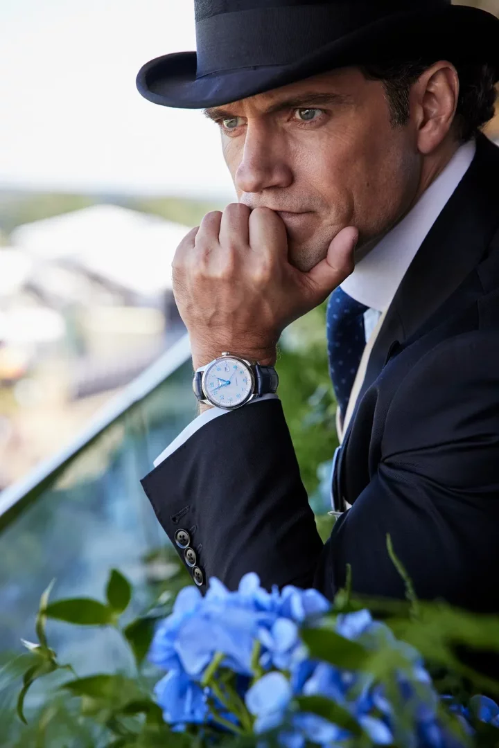 Royal Ascot gibi resmi bir at yarışı etkinliğinin atmosferini yansıtan dış mekan bir balkonda, Henry Cavill siyah silindir şapkası, lacivert takım elbisesi ve kravatıyla, elini çenesine dayamış, düşünceli ve ciddi bir ifadeyle uzaktaki manzaraya bakarken, sol bileğinde beyaz kadranlı ve mavi deri kayışlı zarif bir Longines kol saati dikkat çekiyor.