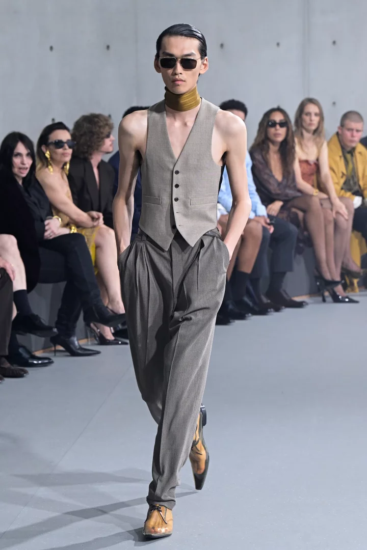 Podyumda kararlı adımlarla yürüyen uzun ve ince erkek model, Saint Laurent İlkbahar-Yaz 2027 erkek giyim koleksiyonundan kolsuz, açık bej bir yelek, yüksek belli bol kesim gri pantolon, boğazında şık bir fular ve güneş gözlükleriyle hafifleyen terzilik ve bedenin görünürlüğü trendini vurguluyor.