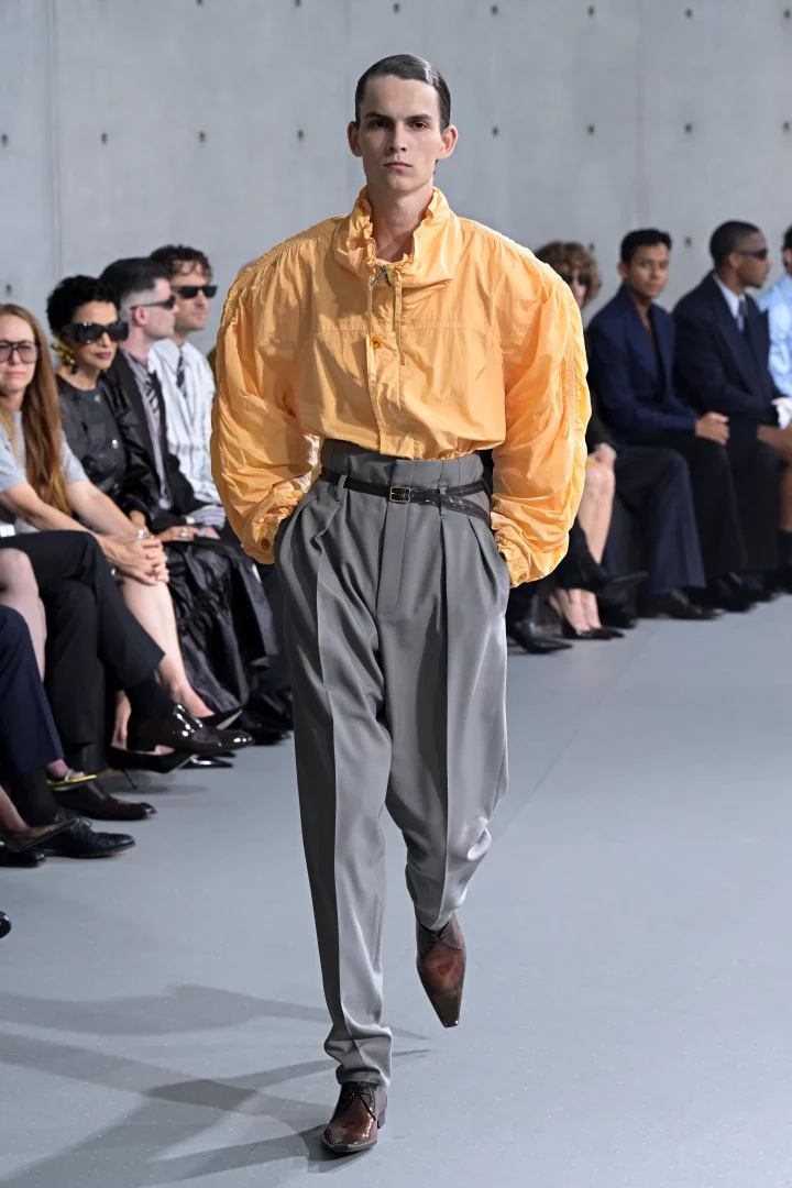 Islak görünümlü saçlara sahip genç bir erkek model, turuncu renkte, bol ve hacimli kollu bir üst ile gri, yüksek belli, bol kesimli, pileli bir pantolonu kombinleyerek Saint Laurent İlkbahar-Yaz 2027 erkek giyim koleksiyonundan modern ve hafifleyen bir terzilik anlayışını podyumda sunuyor.