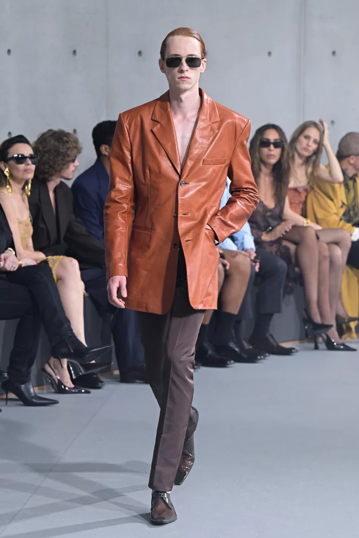 Bir erkek model, Paris İlkbahar-Yaz 2027 Erkek Moda Haftası'nda Saint Laurent defilesinde podyumda yürürken, arkaya taranmış kızılımsı saçları, güneş gözlükleri ve çıplak göğsünü sergileyen pas rengi uzun deri ceketi ile şık kahverengi pantolonunu zarifçe taşıyor.