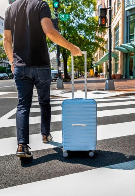 Şehirde güneşli bir günde, yaya geçidinden geçen, sırtı dönük bir adam, modern ve açık mavi bir Samsonite valizini kolayca çekiyor; arka planda yeşil ve kırmızı trafik ışıklarıyla birlikte şehir binaları ve yeşil ağaçlar seçiliyor.