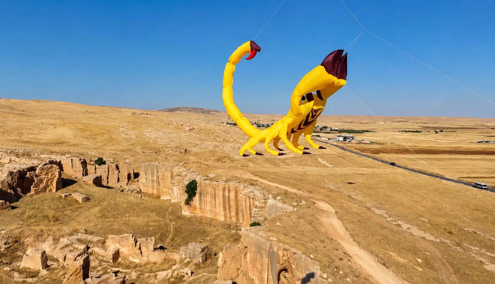 Geniş, kurak bir ovada, üzerinde eski taş yapıların ve uzak bir yolun görüldüğü manzaranın fonunda, kuyruğu kırmızı bir iğneyle biten ve koyu renkli kıskaçlara sahip devasa bir sarı akrep şeklindeki şişme uçurtma, Mardin Bienali'nin sembolik bir eseri olarak masmavi gökyüzünde süzülüyor.