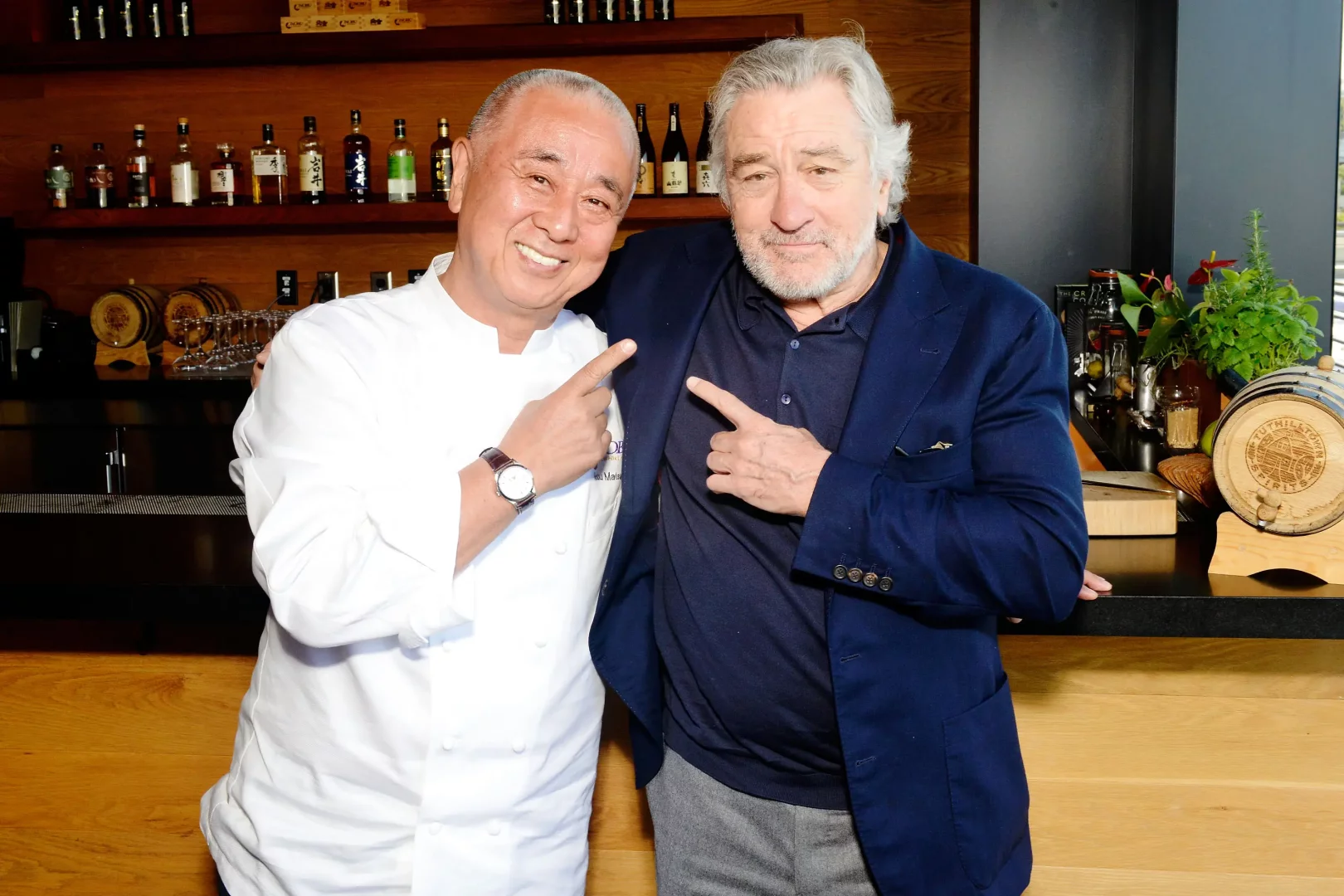 Dünyaca ünlü Şef Nobu Matsuhisa ve ortağı aktör Robert De Niro, Nobu restoranlarının başarısını ve ortaklıklarını vurgularcasına birbirlerini işaret ederek gülümsüyorlar; arkalarında ise markanın global mutfak felsefesini simgeleyen bir bar ve içki şişeleri seçkisi görülüyor.