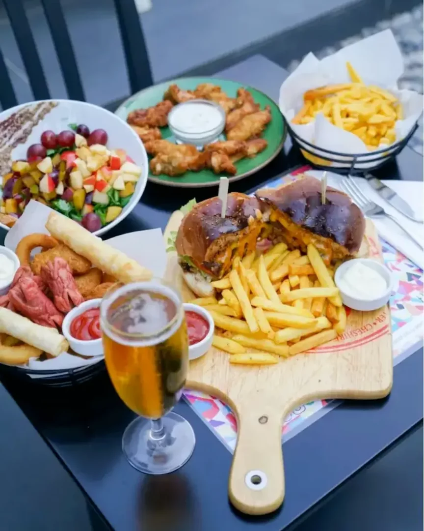 Büyükada'daki Sermest Restoran'da çekilen bu karede, masada iştah açıcı bir hamburger patates kızartmasıyla, çıtır tavuk kanatları, taze meyve salatası, soğan halkaları ve soğuk bir bira eşliğinde zengin bir menü sunumu göze çarpıyor.