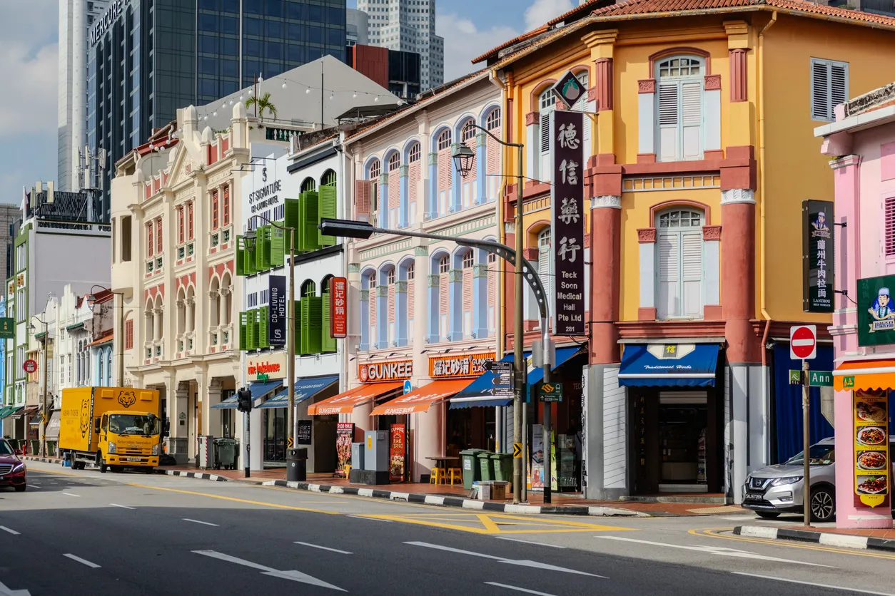 Singapur'un hareketli caddelerinden birinde, geleneksel mimariye sahip, kemerli pencereleri ve süslü cepheleriyle dikkat çeken sıralı, çok renkli dükkan binalarının bulunduğu, arka planda modern gökdelenlerin yükseldiği, tarih ve çağdaşlığın iç içe geçtiği özel bir şehir manzarası.