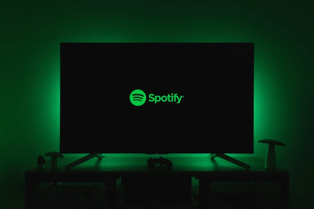Karanlık bir odada, yeşil arka ışıkla aydınlatılmış bir duvara yaslanmış akıllı televizyonun siyah ekranında parlak yeşil Spotify logosu ve adı belirgin bir şekilde görünüyor; televizyonun altındaki sehpada bir oyun kumandası ve dekoratif mantar objeleri seçiliyor.