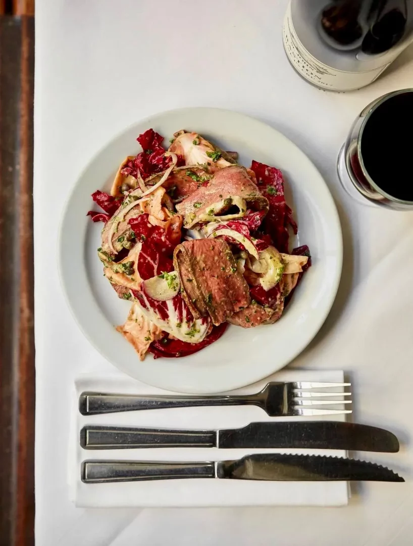 Beyaz masa örtüsü üzerinde, ortasında dilimlenmiş orta az pişmiş et, parlak kırmızı radicchio, ince halka soğanlar ve yeşil sosla zenginleştirilmiş iştah açıcı bir salatanın yer aldığı beyaz bir tabak, yanında dolu bir kırmızı şarap kadehi, bir şarap şişesi ve beyaz peçete üzerine özenle yerleştirilmiş metal çatal ve iki bıçakla şık bir akşam yemeği sahnesi.