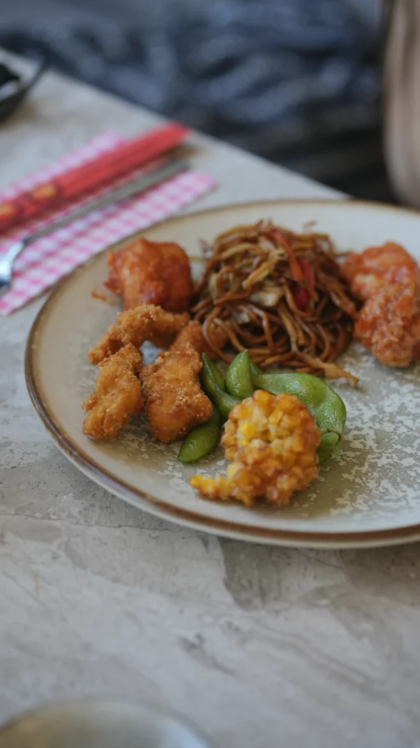 Kızarmış tavuk parçaları, tatlı ekşi soslu büyük karides veya tavuklar, edamame fasulyesi, sotelenmiş erişte ve çıtır mısır tempurası içeren, Asya mutfağının zenginliğini yansıtan dolu dolu bir tabak gri mermer bir masanın üzerinde önden görülüyor.
