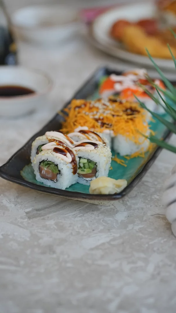Siyah uzun bir tabakta sunulan, önde somon ve salatalık dolgulu, susam serpilmiş, kremalı ve kahverengi soslarla zenginleştirilmiş altı adet uramaki sushi rulosu, yanında bir tutam turşulu zencefil ve arkada çıtır turuncu ipliklerle süslenmiş, farklı tatlar sunan diğer sushi çeşitleri, odak dışında bir kase soya sosuyla birlikte gri, mermer desenli bir masa üzerinde sergileniyor.