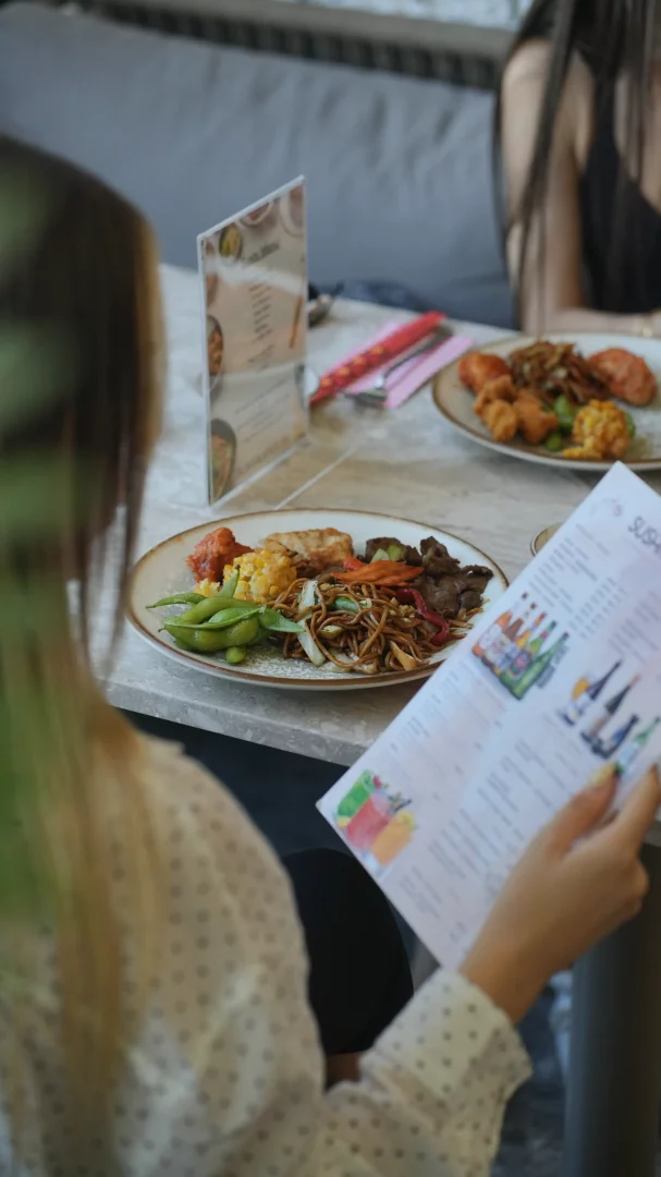 Bir restoranda, omzundan görünen bir kadın elinde Sushi yazılı bir menüye bakarken, masada erişte, edamame, mısır ve çeşitli sebzelerle hazırlanmış Asya mutfağına özgü baharatlı lezzetler dolu zengin bir tabak ve arka planda başka yemekler belirginleşiyor.