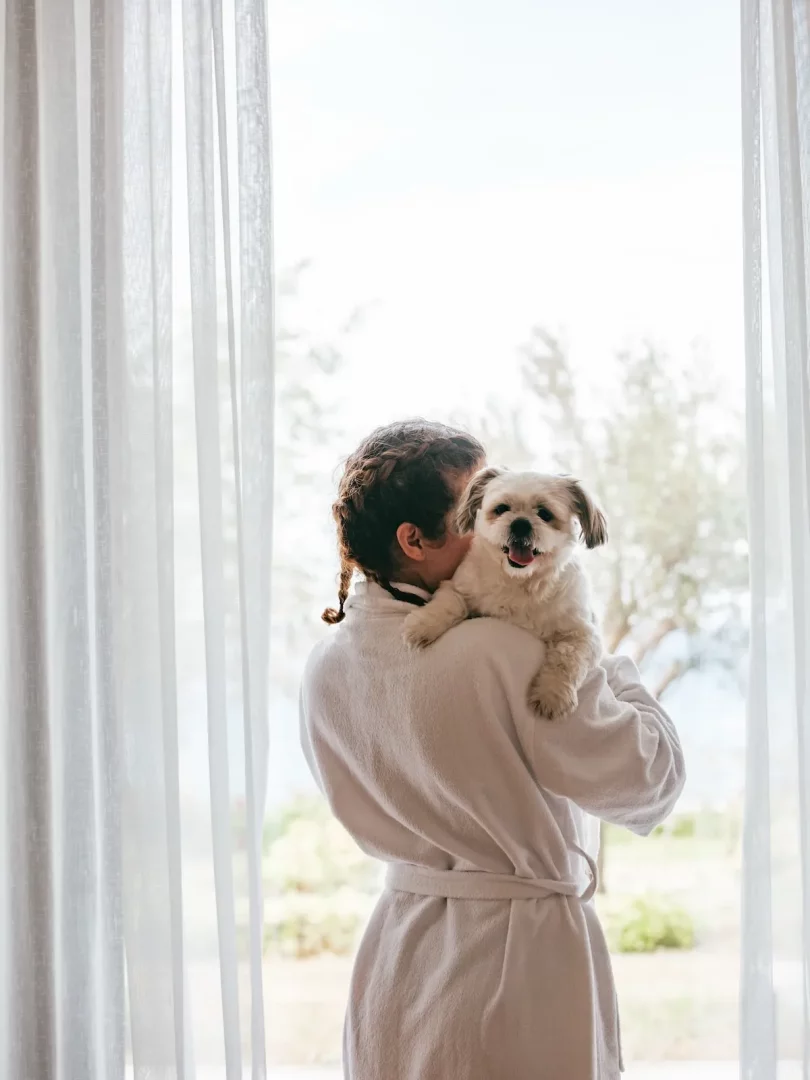 Susona Bodrum'un lüks evcil hayvan dostu konaklama imkanlarını öne çıkaran bu görselde, beyaz bornozlu bir misafir omuzunda sevimli, tüylü beyaz köpeğiyle pencereden dışarıdaki yeşil manzarayı seyrediyor.