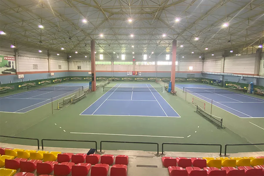 TAC Spor Kulübü'ne ait geniş iç mekan tenis kortlarından bir görünüm; parlak ışıklarla aydınlatılmış, mavi zeminli çok sayıda kort, tribünler ve spor kompleksinin yüksek tavanlı yapısı dikkat çekiyor.