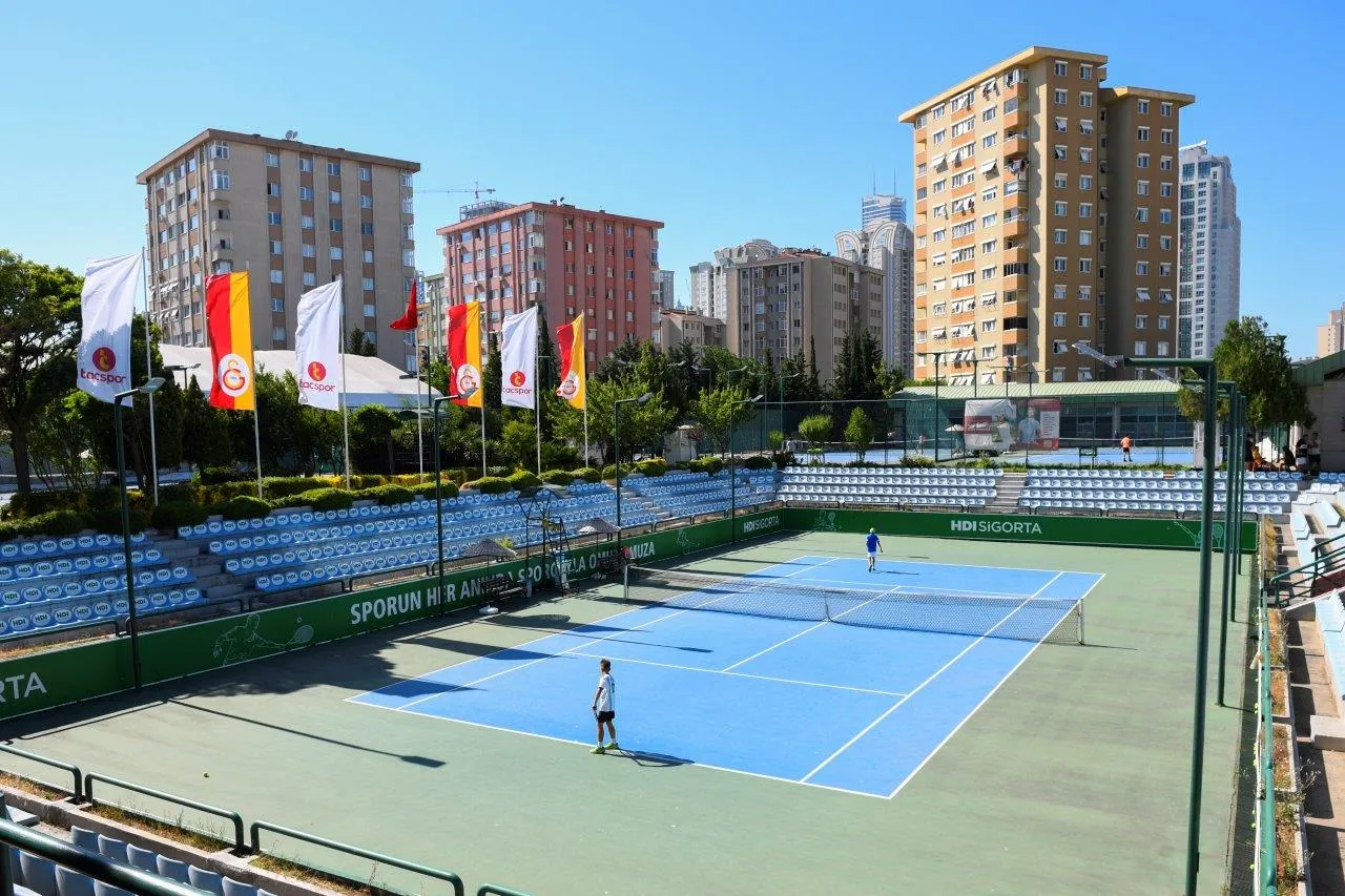 TAC Spor Kulübü'nün açık mavi tenis kortu, üzerinde oynayan iki kişiyle, çevresindeki boş seyirci tribünleri ve şehir silüetindeki çok katlı binalarla birlikte, Tacspor ile Galatasaray bayraklarının dalgalandığı güneşli bir ortamda geniş açıyla gösterilmektedir.