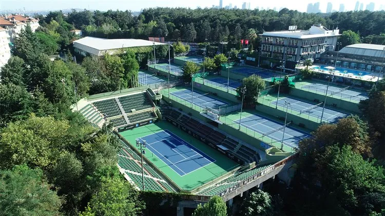TED Spor Kulübü'nün yemyeşil ağaçlarla çevrili geniş yerleşkesini kuşbakışı gösteren bu fotoğrafta, seyirci tribünleriyle dikkat çeken ana tenis kortu ve farklı seviyelere yerleştirilmiş, spor kompleksi binalarının da görüldüğü çok sayıda mavi zeminli tenis kortu yer almaktadır.