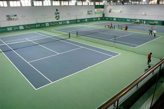 TED Spor Kulübü'nün geniş kapalı tenis kortu kompleksinde, parlak ışıklar altında yeşil zemin üzerine kurulu birkaç mavi tenis kortunda, insanlar tenis oynuyor, yürüyor ve sosyalleşiyor; arka planda kulübün logosu ve skorbordu da görülüyor.