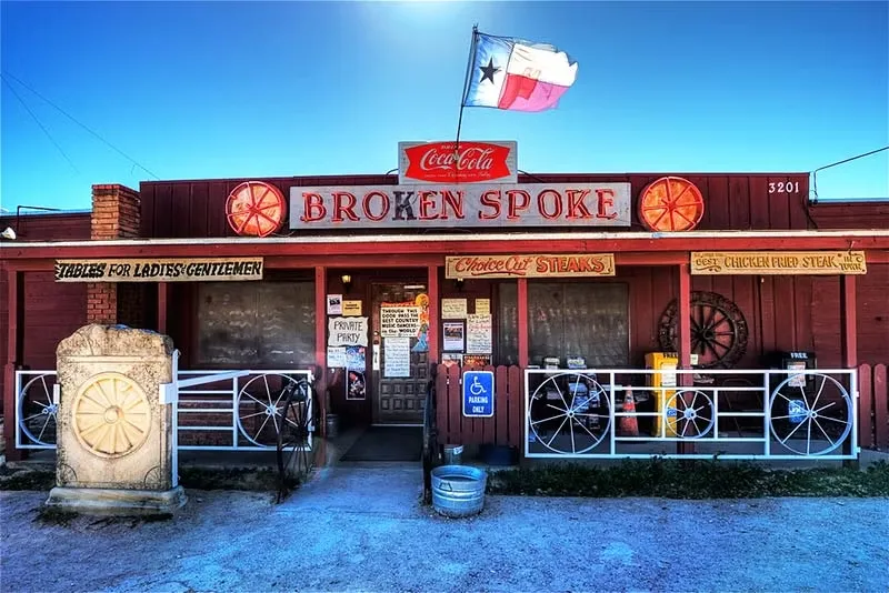 Güneşli mavi gökyüzü altında Teksas bayrağının dalgalandığı, batı temalı kırmızı cephesiyle ikonik Broken Spoke lokantasının dış görünüşü; menüsünde biftek ve kızarmış tavuk gibi yemekleri belirten tabelalar, girişinde eski tekerlek motifleri ve engelli park yeri işaretiyle detaylı bir şekilde gösteriliyor.