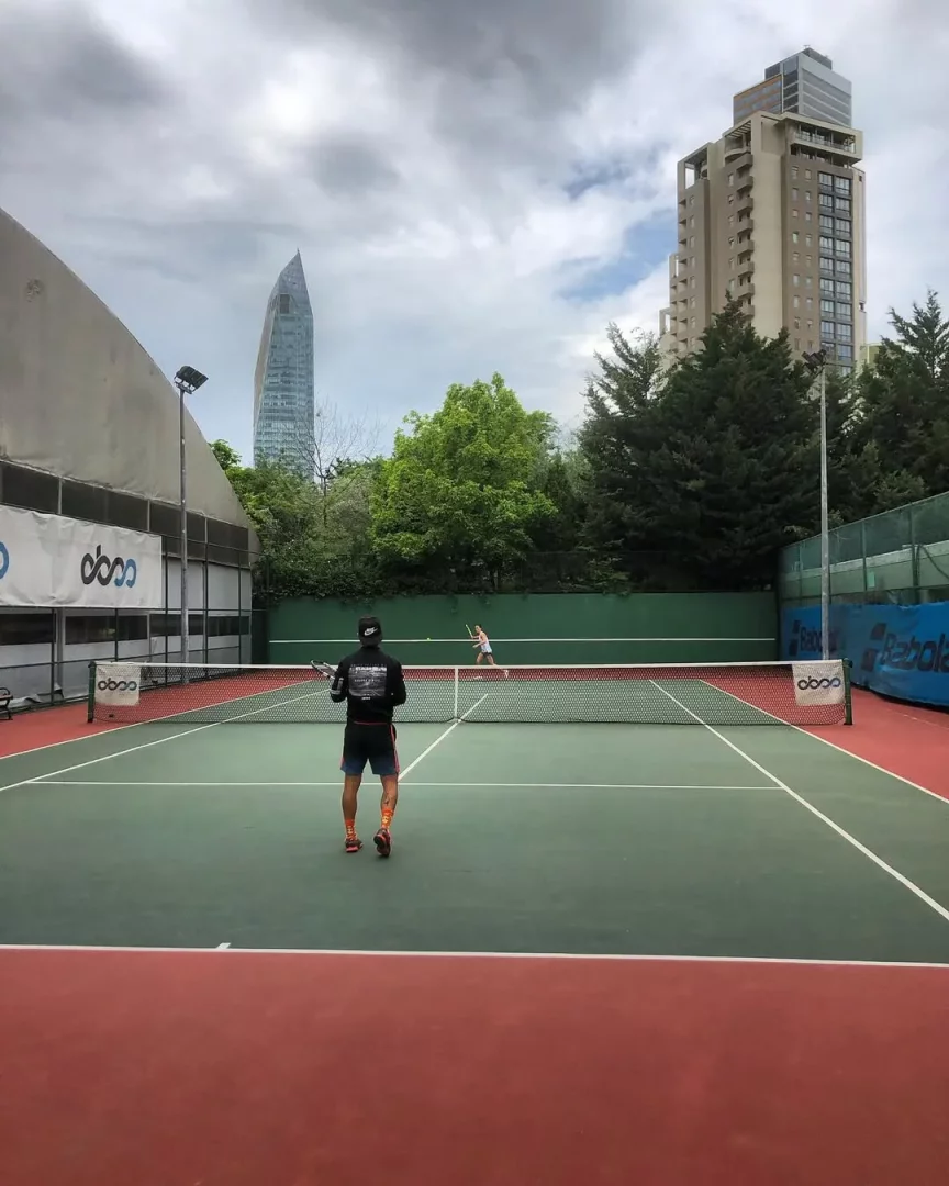 Ataşehir'deki bir tenis kortunda, bulutlu gökyüzü altında iki kişinin yemyeşil sahada tenis oynadığı, ön planda sırtı dönük raketli bir oyuncunun ve arka planda İstanbul'un modern yüksek binalarının göründüğü geniş bir manzara.