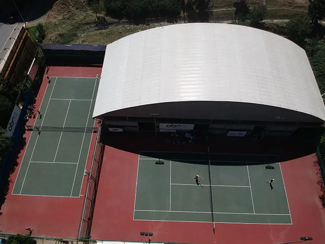 Ataşehir Tenis Kulübü'nün geniş açıdan çekilmiş bu fotoğrafında, kırmızı zeminli alana yayılmış yemyeşil kortlarda beyaz çizgilerle belirlenmiş oyun alanları ve kortların bir bölümünü kaplayan büyük, modern, beyaz kemerli çatı yapısı dikkat çekiyor, bazı kortlarda oyuncular tenis oynuyor.