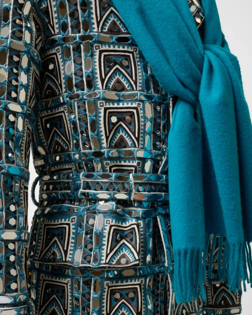 Büyüleyici aynalı ve nakışlı geometrik desenlere sahip, teal, siyah ve beyaz tonlardaki kemerli bir haute couture giysi, üzerine zarifçe atılmış püsküllü düz teal bir atkıyla sergilenerek, modanın zanaatla buluştuğu 2026 yılındaki önemli moda sergilerinin görsel bir özeti gibi duruyor.