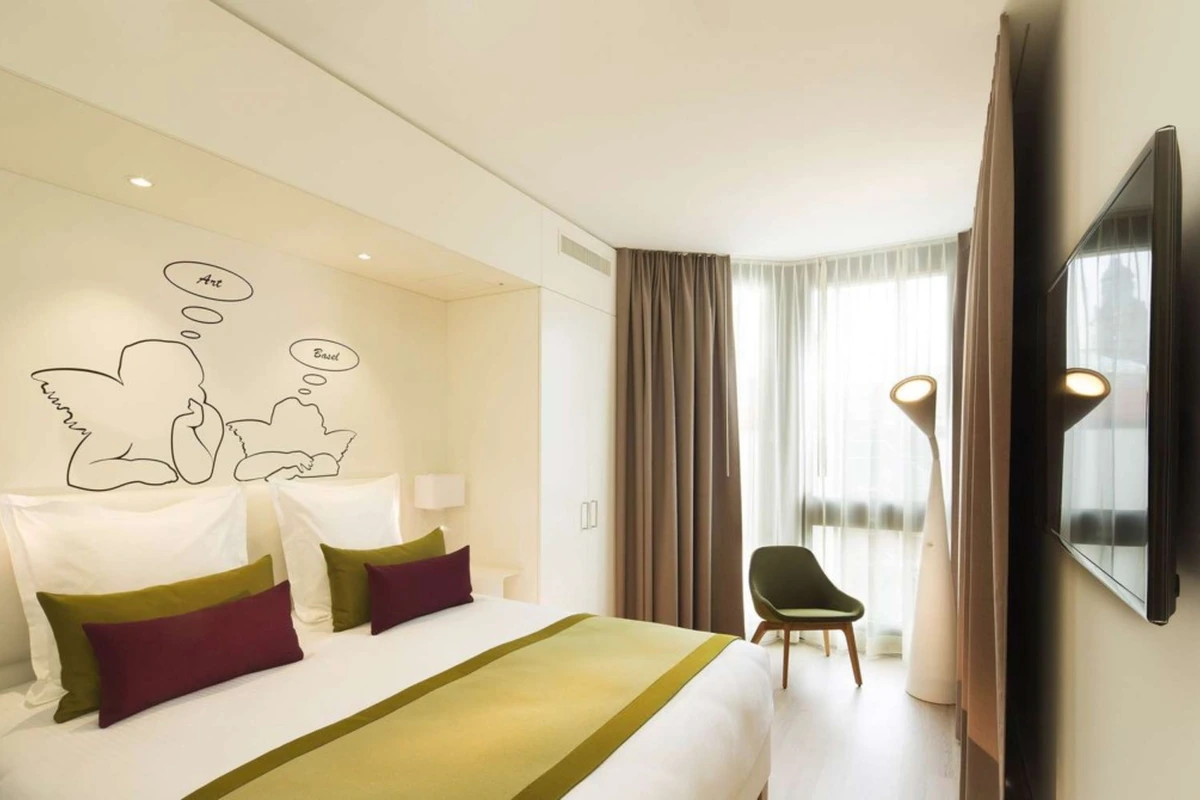 The Passage Urban Retreat oteline ait bu modern odada, beyaz çarşaflı ve yeşil-bordo yastıklı yatağın başucunda Art ve Basel yazılı iki melek figürü, sağ tarafta tül ve koyu perdeli büyük bir pencere önünde yeşil bir sandalye ve dekoratif zemin lambası, sağ duvarda ise duvara monte bir televizyon ile ferah ve aydınlık bir yaşam alanı görülüyor.