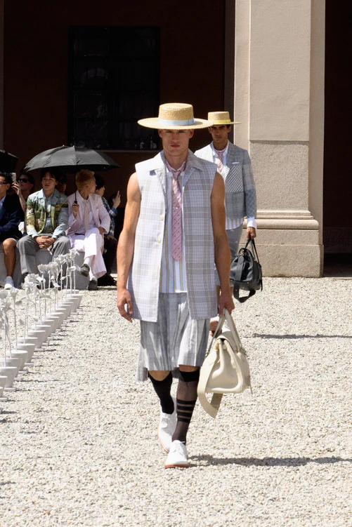 Thom Browne'ın Milano'daki İlkbahar-Yaz 2027 erkek giyim defilesinden bir model, açık havada, kolsuz ekose bir yelek, çizgili bir gömlek, pembe kravat, pilili kısa etek, dizüstü çoraplar ve hasır şapka giyerek yürüyor, beyaz bir el çantası taşırken, arkasında benzer giysili başka bir model ve izleyicilerle dolu 'Thom's Garden' temalı bir podyumda Amerikan prep estetiğini sergiliyor.