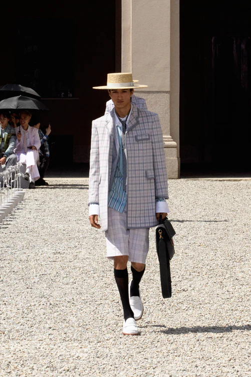 Thom Browne'ın İlkbahar-Yaz 2027 erkek giyim koleksiyonundan, hafifleyen terzilik ve yeni Amerikan zarafetini yansıtan; ekoseli uzun blazer ceket, uyumlu kısa şort, çizgili gömlek, kravat, hasır şapka, siyah çorap, beyaz makosen ayakkabılar ve siyah evrak çantası giyen bir model çakıllı podyumda ilerliyor.