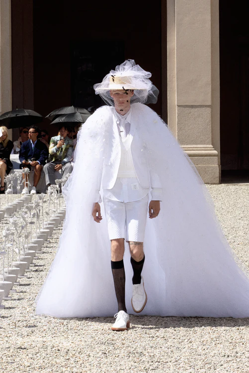 Thom Browne'un Milano Moda Haftası'ndaki İlkbahar-Yaz 2027 erkek giyim koleksiyonundan, arı motifli tüllü şapka ve görkemli pelerinle tamamlanan beyaz kısa takım elbiseli bir model, defile alanında taşlı yolda yürüyor.