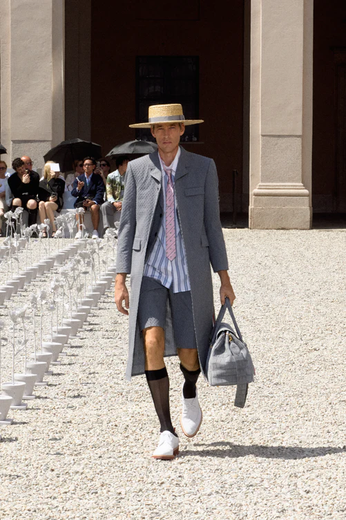 Milano Erkek Moda Haftası İlkbahar-Yaz 2027 defilesinde, gri seersucker kumaştan uzun bir palto, şort, çizgili gömlek ve pembe kravat giyen, hasır şapkalı bir model beyaz çiçeklerle süslü podyumda, bahçe temasına uygun şık ve rahat erkek giyim tarzını sergiliyor.