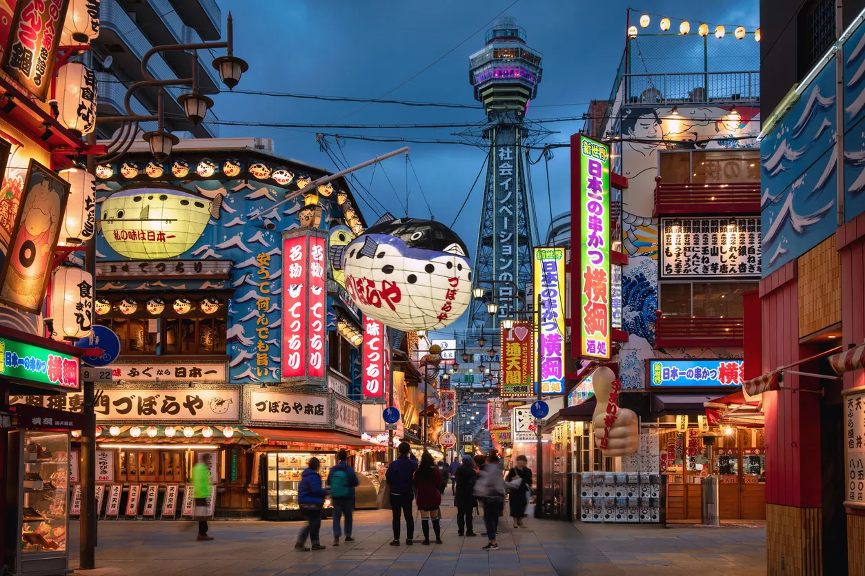 Osaka'nın Shinsekai bölgesinde, akşam alacakaranlığında neon ışıklı reklam panoları, devasa kirpi balığı fenerleri ve arka plandaki ikonik Tsutenkaku Kulesi'nin aydınlattığı kalabalık bir sokakta insanlar yürüyor.