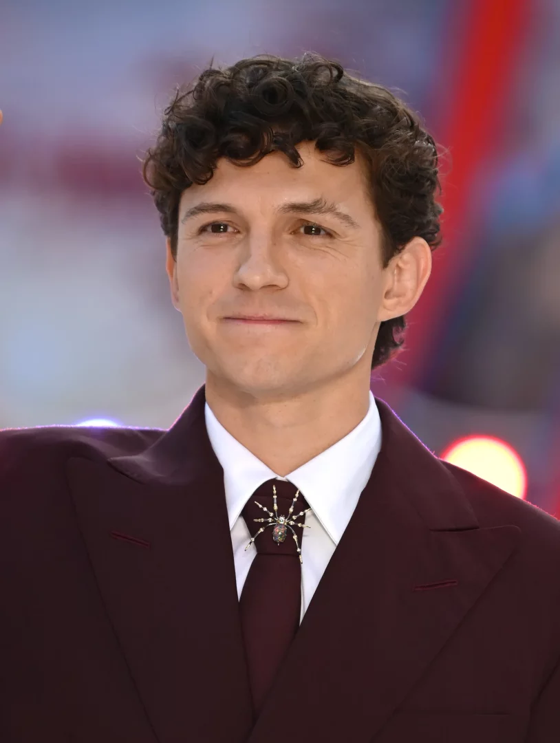 Tom Holland, Örümcek-Adam prömiyerinde hafifçe gülümserken, kıvırcık saçları ve bordo bir takım elbise içinde görülüyor; beyaz gömleği ve bordo kravatının düğümünü süsleyen, dikkat çekici, detaylı bir örümcek broş karakterine gönderme yapıyor.