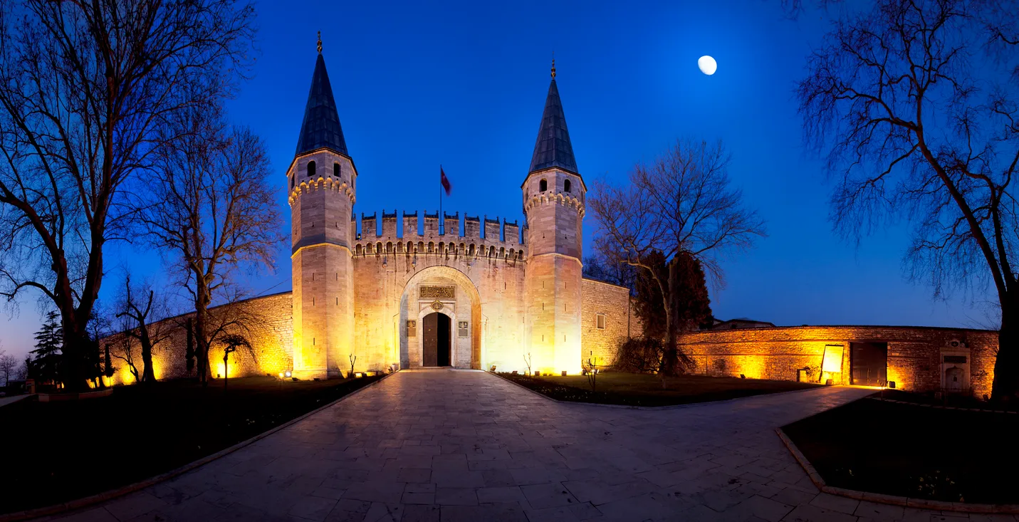 Gece müzesi olarak aydınlatılan Topkapı Sarayı'nın iki kuleli ana giriş kapısı, sol üstte beliren dolunay ve etrafındaki çıplak ağaçlar eşliğinde göz alıcı bir manzara oluşturuyor.