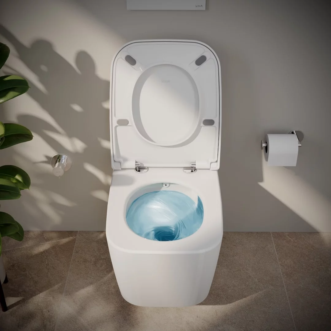 Beyaz, modern tasarımlı bir Vitra klozet, açık kapak ve oturağıyla, içinde güçlü ve hijyenik bir temizliği gösteren dairesel mavi su girdabıyla, sağında tuvalet kağıdı ve solunda bitki gölgesinin düştüğü açık renkli duvara karşı konumlanmış, QuantumFlush sisteminin verimliliğini sergiliyor.