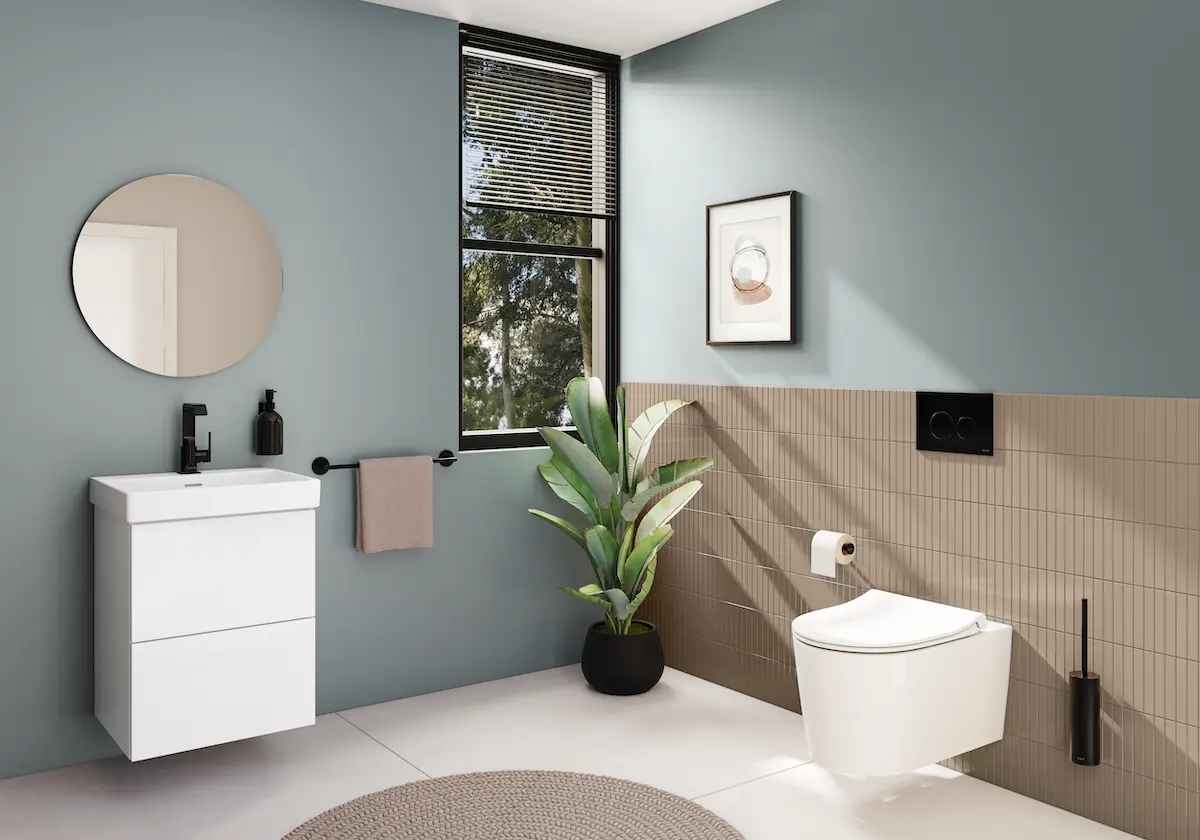 Modern ve şık bir banyo düzenlemesi, VitrA QuantumFlush asma klozetin öne çıktığı, açık mavi duvarlar ve bej dikey fayanslarla kaplı bir alanda, yuvarlak ayna, beyaz dolaplı lavabo, siyah armatürler, büyük bir bitki ve doğal ışıkla aydınlatılmış ferah bir ambiyans sunuyor.