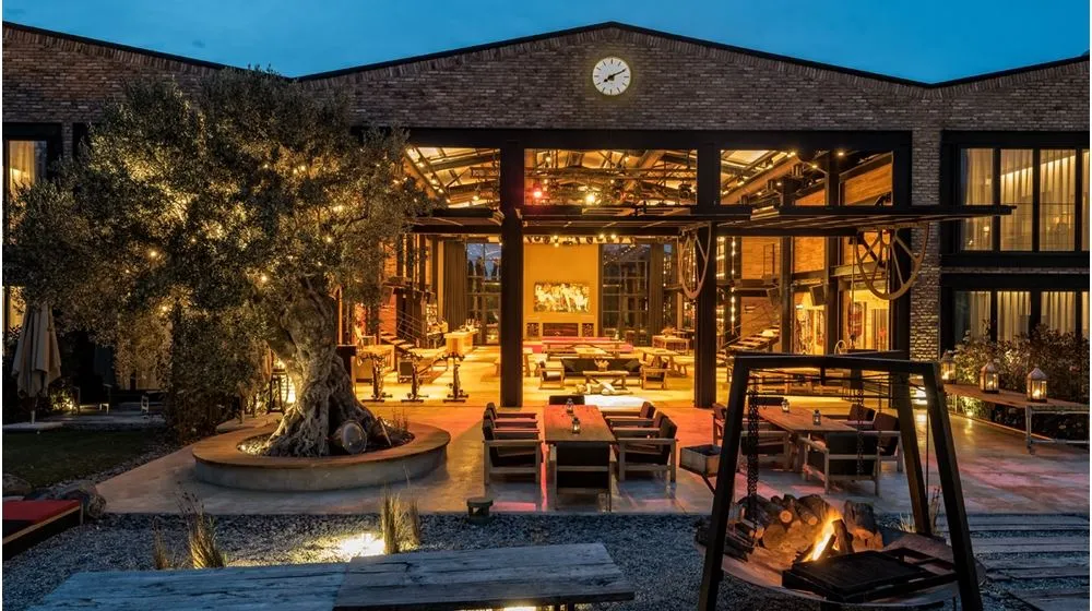 Bir depodan dönüştürülmüş modern otel girişi, dışarıda ışıklandırılmış zeytin ağacı, oturma alanları ve yanan bir ateş çukuru ile sıcak bir akşam ambiyansı sunarken, içeride geniş ve şık bir lobi alanı görülüyor.
