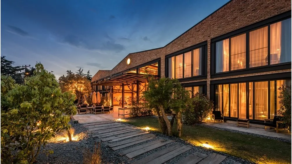 Bir zamanlar depo olan The Stay otelinin tuğla dış cephesi ve içeriden sıcak ışıklarla aydınlanan geniş pencereleriyle modern mimarisi, ahşap yürüme yolu, bakımlı bahçesi ve dış mekan oturma alanları, alacakaranlıkta loş ışıklarla çevrili huzurlu bir atmosfer sunuyor.
