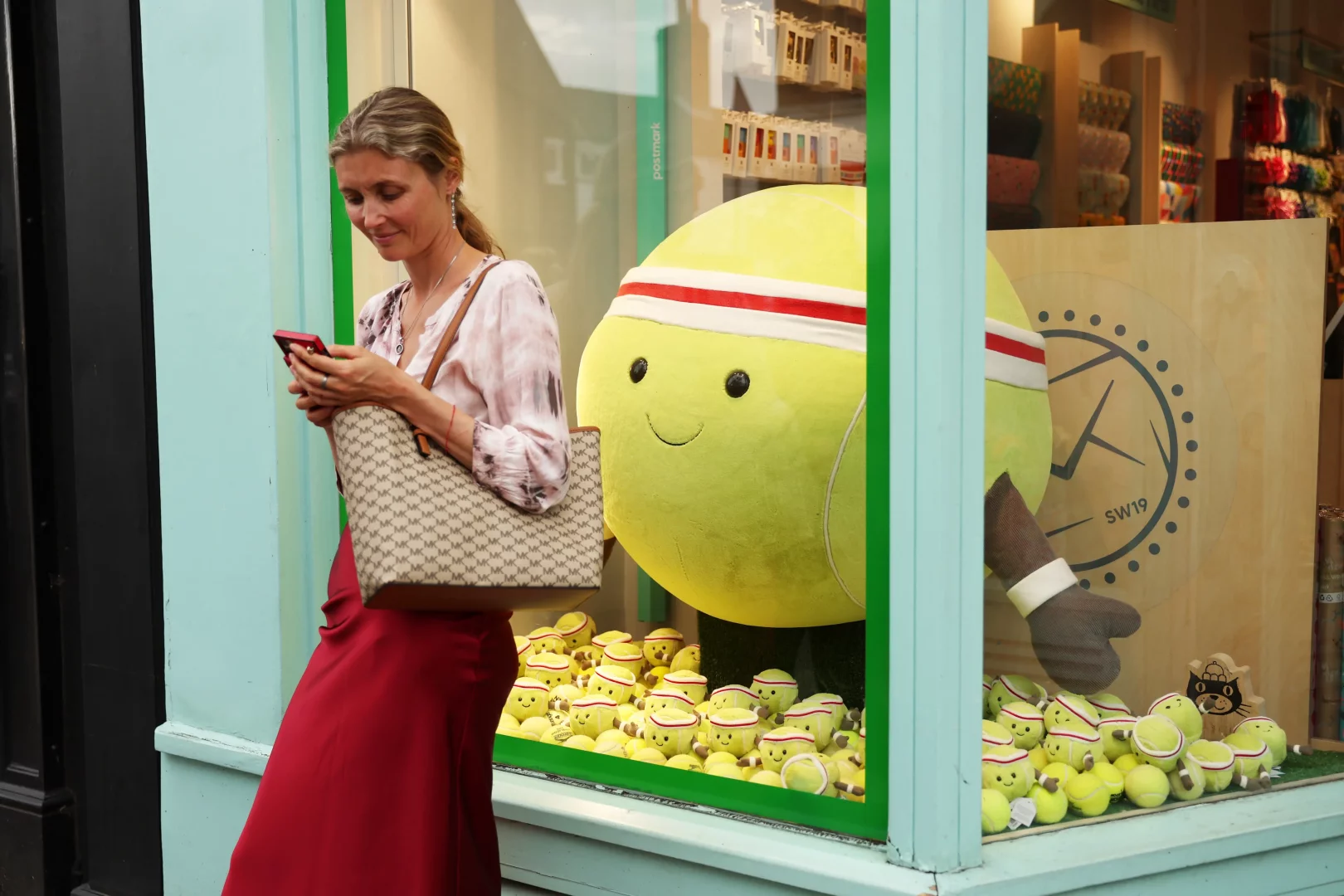 Açık renk bluzu ve kırmızı uzun eteğiyle Wimbledon Village'da şık görünen bir kadın, monogram desenli çantasını tutarak telefonuna bakarken, kırmızı-beyaz kafa bandı takmış gülen büyük bir pelüş tenis topu ve çok sayıda küçük sevimli tenis topu oyuncağıyla dolu, Wimbledon temalı bir butik mağazanın vitrininin önünde duruyor.