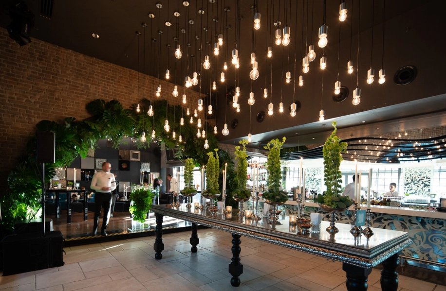 Wyndham Ankara'nın modern ve çok yönlü etkinlik alanlarından birinde, koyu tavanlardan sarkan çok sayıda parlak ampulün aydınlattığı, işlemeli uzun bir masanın üzerinde yükselen yeşil bitki aranjmanları ve gümüş şamdanlar zarif bir düğün veya davet ortamı sunuyor; tuğla duvarlar ve yoğun yeşilliklerle çevrili mekanda, arka planda servis hazırlığı yapan personel ve şık bar alanı da görülmekte.