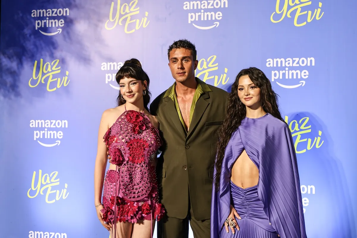 Yaz Evi dizisinin gala etkinliğinde, Amazon Prime ve Yaz Evi logolu mavi fonun önünde poz veren başrol oyuncuları; pembe kroşe detaylı elbisesiyle bir kadın, zeytin yeşili takım elbisesiyle bir erkek ve lavanta rengi dekolteli pilili elbisesiyle diğer bir kadın gülümseyerek kameraya bakıyor.